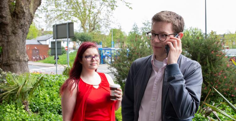 A man is on the phone and the woman next to him is looking at him and holding a hot drink.