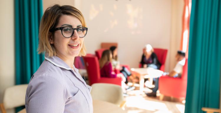 Nurse smiling at the camera. People in the background are sitting round a table chatting. 