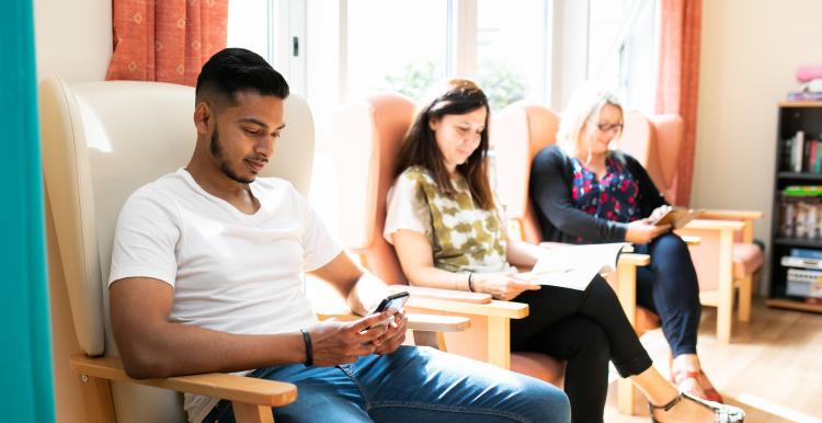 Three people sitting in an hospital waiting room, keeping themselves occupied with magazines and their smartphones