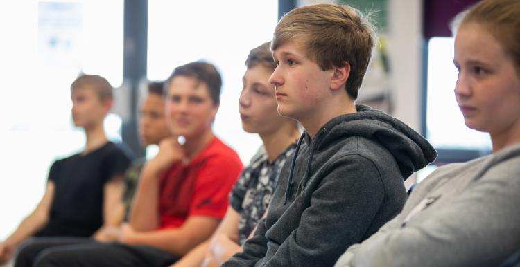 A teenage boy sitting in a row of chairs with other teenagers his age. 