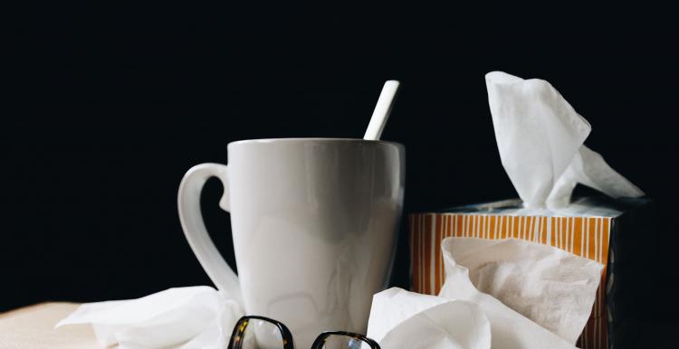 A stack of used tissues on a bedside table, next to a hot cuppa and some folded up glasses. 
