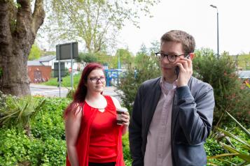 A man is on the phone and the woman next to him is looking at him and holding a hot drink. 