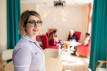 Nurse smiling at the camera. People in the background are sitting round a table chatting. 