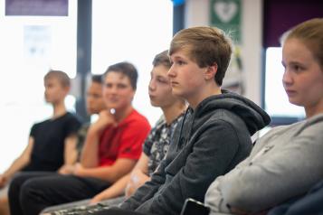 A teenage boy sitting in a row of chairs with other teenagers his age. 