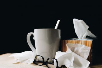 A stack of used tissues on a bedside table, next to a hot cuppa and some folded up glasses. 