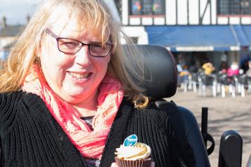 A woman in a wheelchair smiling at the camera, holding a vanilla cupcake with frosting.